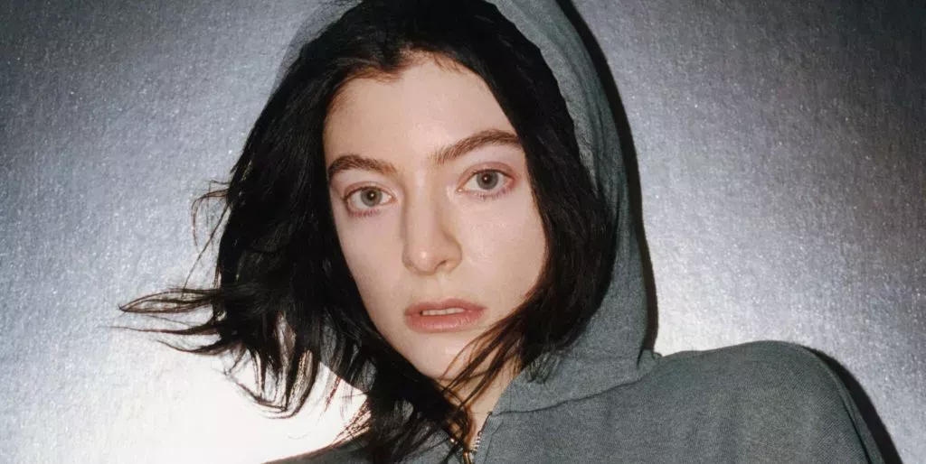 Lorde en una sudadera gris con el cabello castaño asomando mientras está de pie contra un fondo gris.