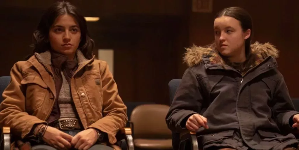 A still from episode one of The Last of Us featuring characters Dina and Ellie sitting on chairs, with Ellie looking over at Dina scornfully.